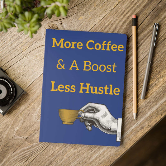 Coffee-Themed Softcover Journal with "More Coffee & A Boost Less Hustle" text on blue cover alongside pencil and pen on wooden table