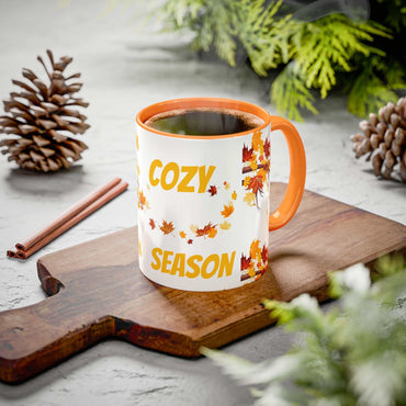 Fall Cozy Season Mug with orange handle featuring autumn leaves and "Cozy Season" text, filled with hot coffee on wooden board with pine cones and greenery background