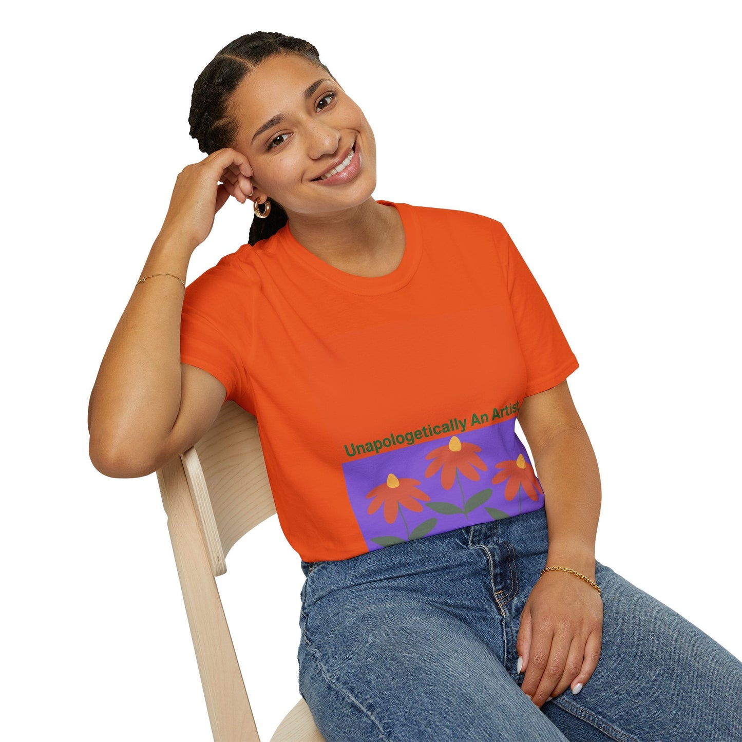 Woman wearing orange Unapologetically An Artist T-Shirt with floral design sitting on chair and smiling