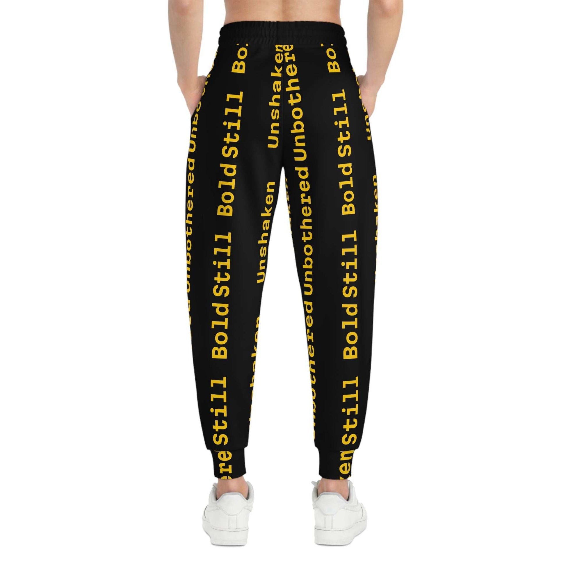 Back view of eco-friendly joggers with black base and bold yellow "Unbothered Unshaken Bold Still" text print, worn with white sneakers