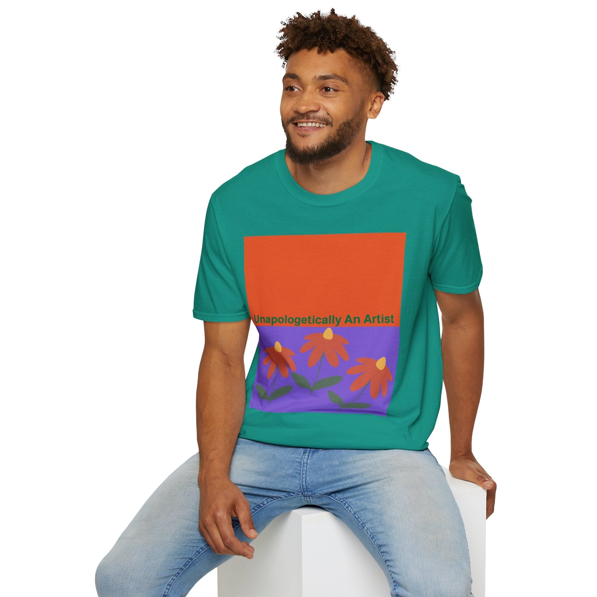 Man wearing green Unapologetically An Artist T-Shirt with floral design and text, blue jeans, sitting on white cube