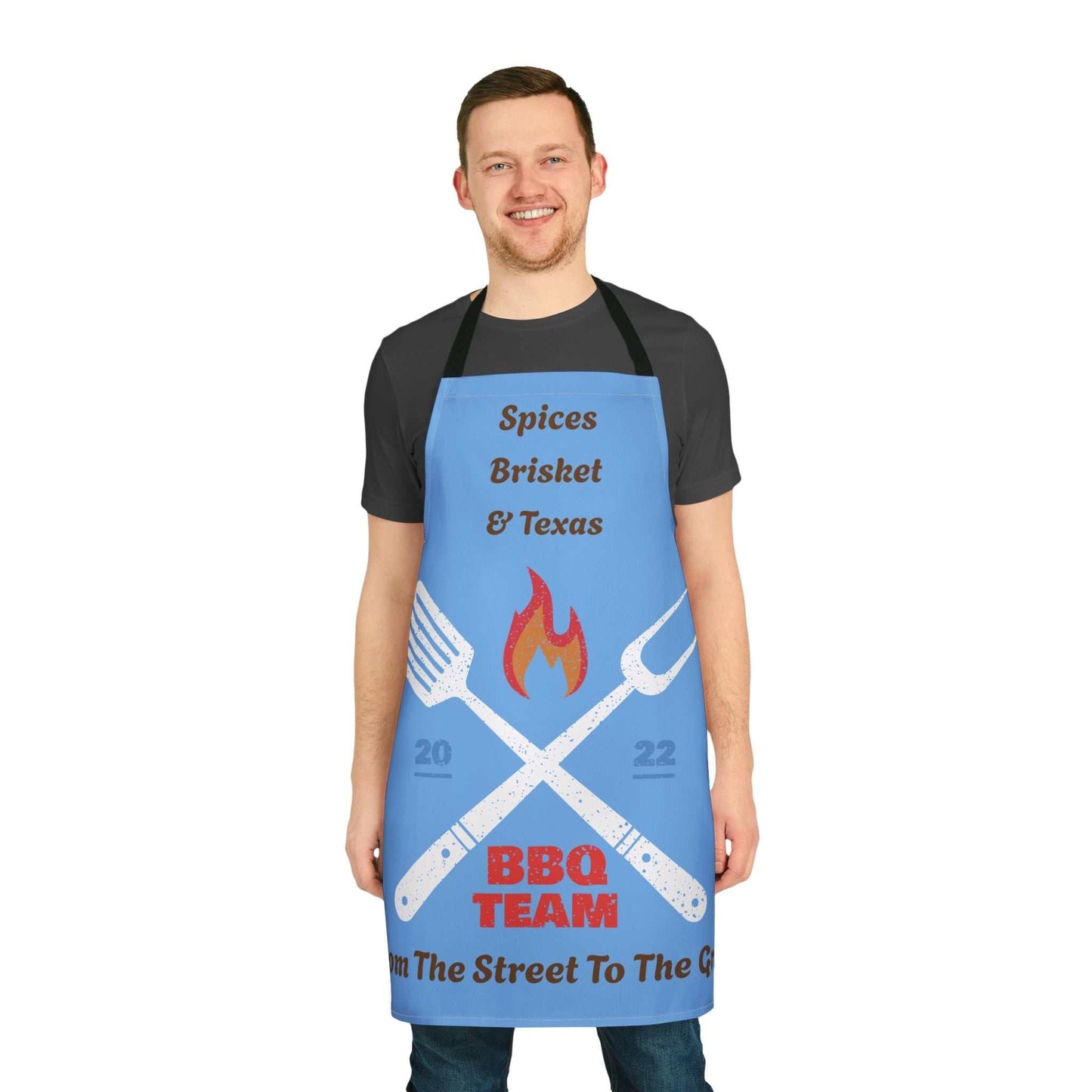 Man wearing blue apron with BBQ team design and black straps standing smiling